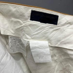 Puede incluir: Una prenda de algod&oacute;n blanco con una cintura marr&oacute;n y una etiqueta negra que dice "COMME des GARCONS". La etiqueta est&aacute; cosida en la cintura. La prenda tiene una etiqueta de cuidado con texto japon&eacute;s.