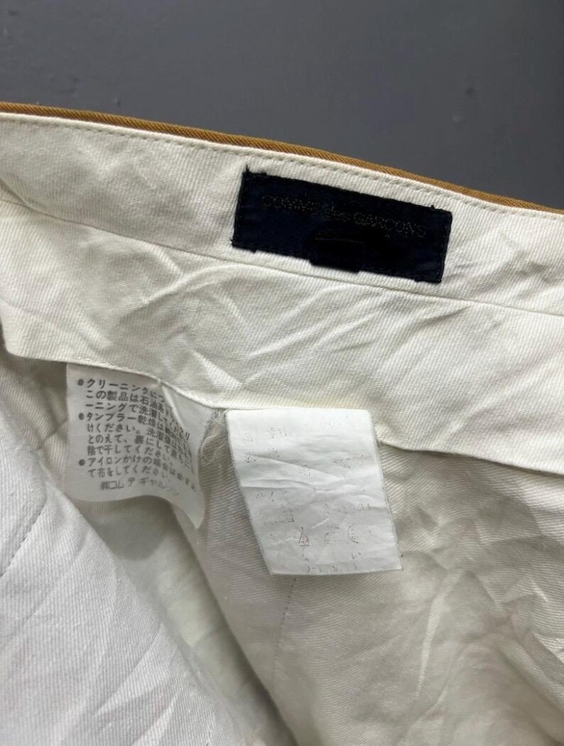 Puede incluir: Una prenda de algod&oacute;n blanco con una cintura marr&oacute;n y una etiqueta negra que dice "COMME des GARCONS". La etiqueta est&aacute; cosida en la cintura. La prenda tiene una etiqueta de cuidado con texto japon&eacute;s.