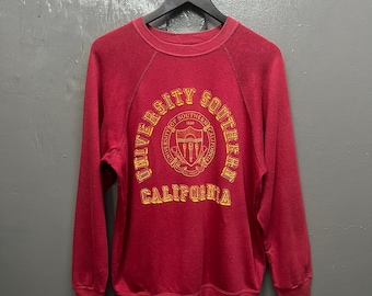 May include: A red crewneck sweatshirt with the words "UNIVERSITY OF SOUTHERN CALIFORNIA" in gold lettering. The sweatshirt has long sleeves and a classic athletic design, perfect for casual wear.