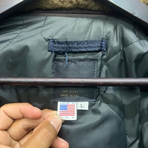 Puede incluir: Primer plano del forro acolchado verde oscuro de una chaqueta. Una etiqueta dice "DRY CLEAN ONLY MADE IN THE USA" y una etiqueta m&aacute;s peque&ntilde;a muestra la bandera estadounidense y "MADE IN USA" con una talla "L".