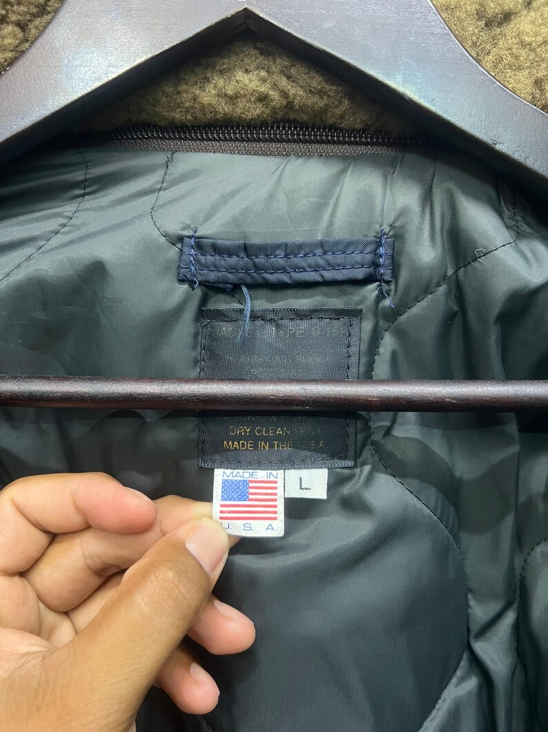 Puede incluir: Primer plano del forro acolchado verde oscuro de una chaqueta. Una etiqueta dice "DRY CLEAN ONLY MADE IN THE USA" y una etiqueta m&aacute;s peque&ntilde;a muestra la bandera estadounidense y "MADE IN USA" con una talla "L".