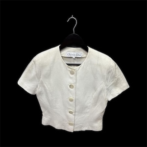 May include: A white, short-sleeved jacket with a textured pattern and a button-up front. The jacket is hanging on a black hanger against a black background. The label reads "Christian Dior".