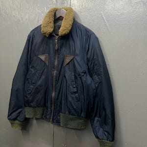 Puede incluir: Chaqueta bomber azul marino con cuello de piel marr&oacute;n y parches de cuero marr&oacute;n. La chaqueta tiene cierre de cremallera y pu&ntilde;os y dobladillo de canal&eacute; verde. La chaqueta est&aacute; colgada de una percha de madera.