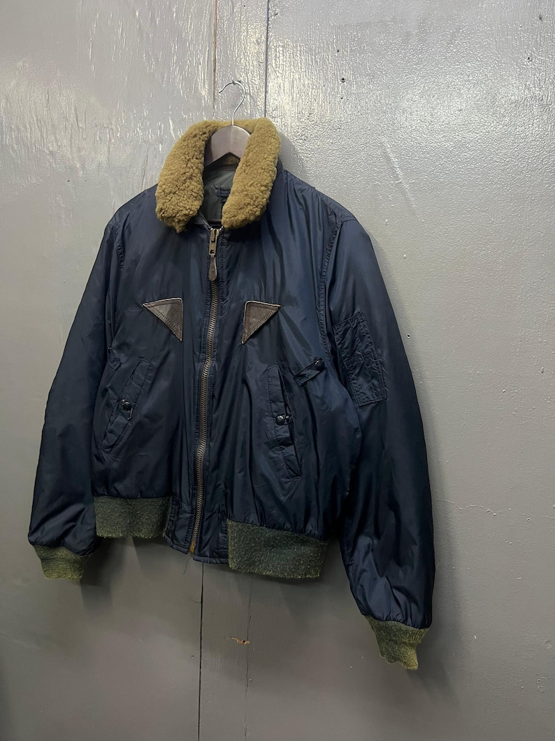 Puede incluir: Chaqueta bomber azul marino con cuello de piel marr&oacute;n y parches de cuero marr&oacute;n. La chaqueta tiene cierre de cremallera y pu&ntilde;os y dobladillo de canal&eacute; verde. La chaqueta est&aacute; colgada de una percha de madera.