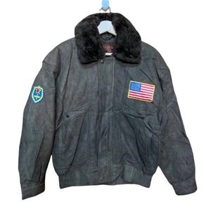 May include: A dark gray leather bomber jacket with a faux fur collar. The jacket has two pockets on the front and a zipper closure. It features an American flag patch and a military-style patch on the left sleeve.