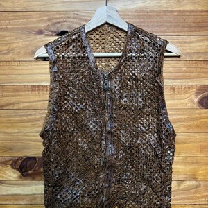 May include: A brown, open-weave vest with a zipper closure. The vest is made of a textured, possibly crocheted or knitted material. It hangs on a wooden hanger against a wooden background.