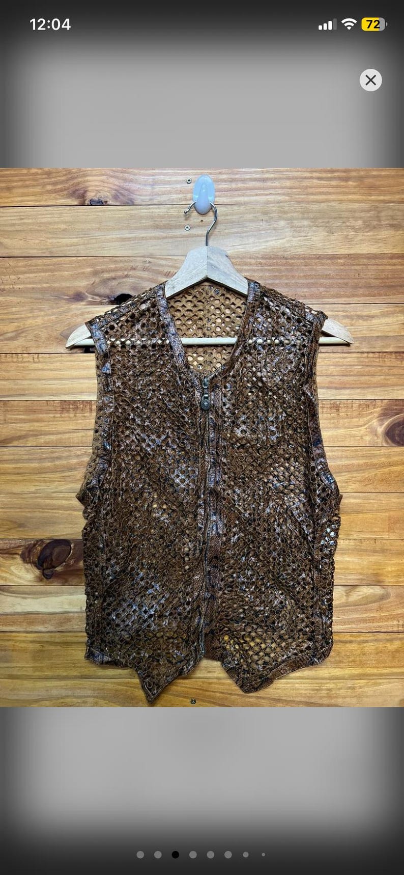 May include: A brown, open-weave vest with a zipper closure. The vest is made of a textured, possibly crocheted or knitted material. It hangs on a wooden hanger against a wooden background.
