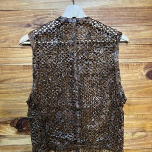 May include: A brown, open-weave vest with a unique, textured pattern. The vest is hanging on a wooden hanger against a wooden background. The vest's design is intricate, with a semi-sheer appearance.