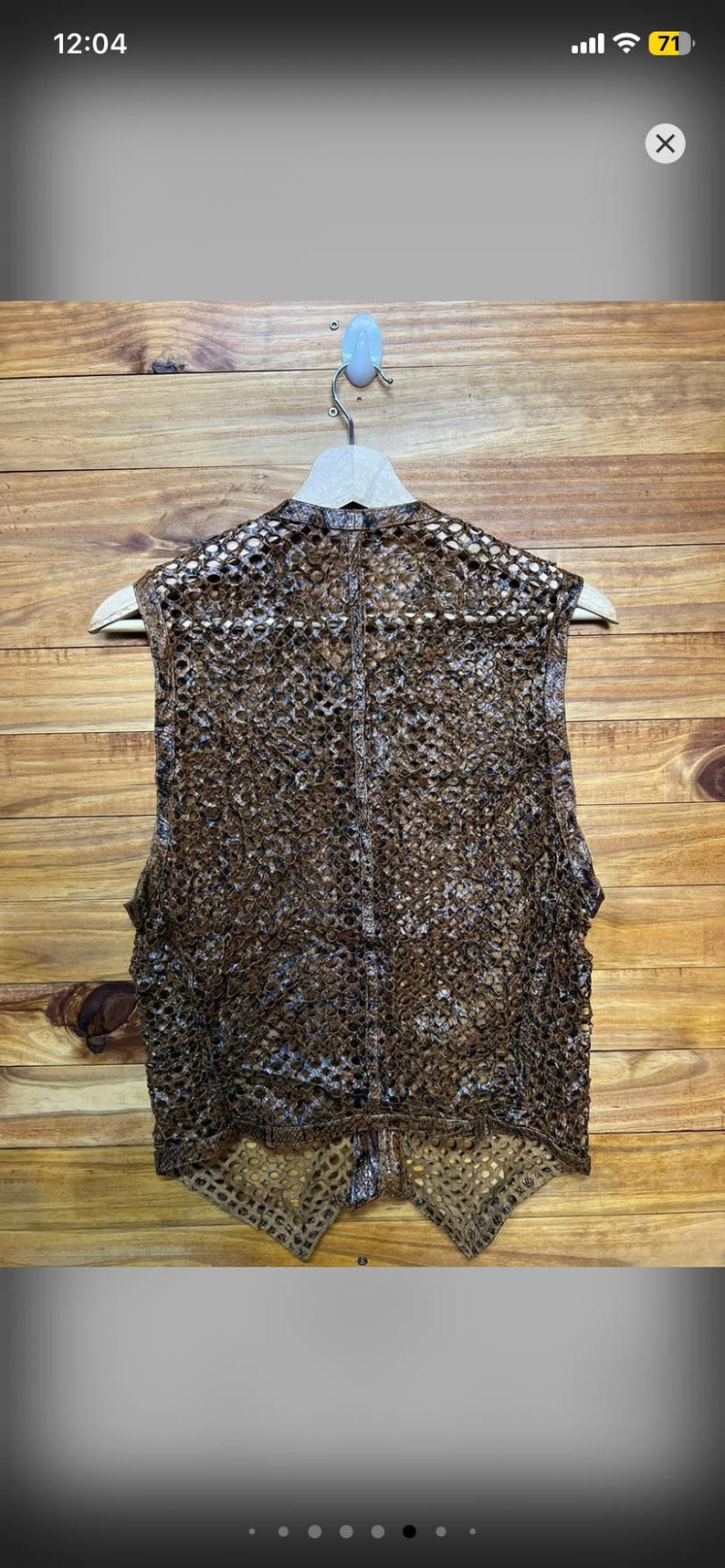 May include: A brown, open-weave vest with a unique, textured pattern. The vest is hanging on a wooden hanger against a wooden background. The vest's design is intricate, with a semi-sheer appearance.