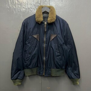 Puede incluir: Una chaqueta bomber azul marino con cuello de borrego marr&oacute;n y parches de cuero marr&oacute;n. La chaqueta tiene una cremallera de lat&oacute;n y pu&ntilde;os y dobladillo de canal&eacute; verdes. La chaqueta est&aacute; colgada en una percha.