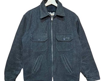 Può includere: Giacca grigio antracite con cerniera a tutta lunghezza e collo a camicia. La giacca ha due tasche sul petto e due tasche inferiori. Il marchio "Schott" è visibile sul petto. La giacca è appesa a un'appendiabiti di legno.