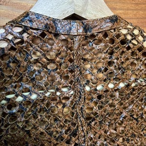May include: Close-up of a brown and black open-weave jacket. The jacket has a unique pattern of cut-out shapes, creating an intricate design. The garment is displayed on a wooden hanger against a wooden background.
