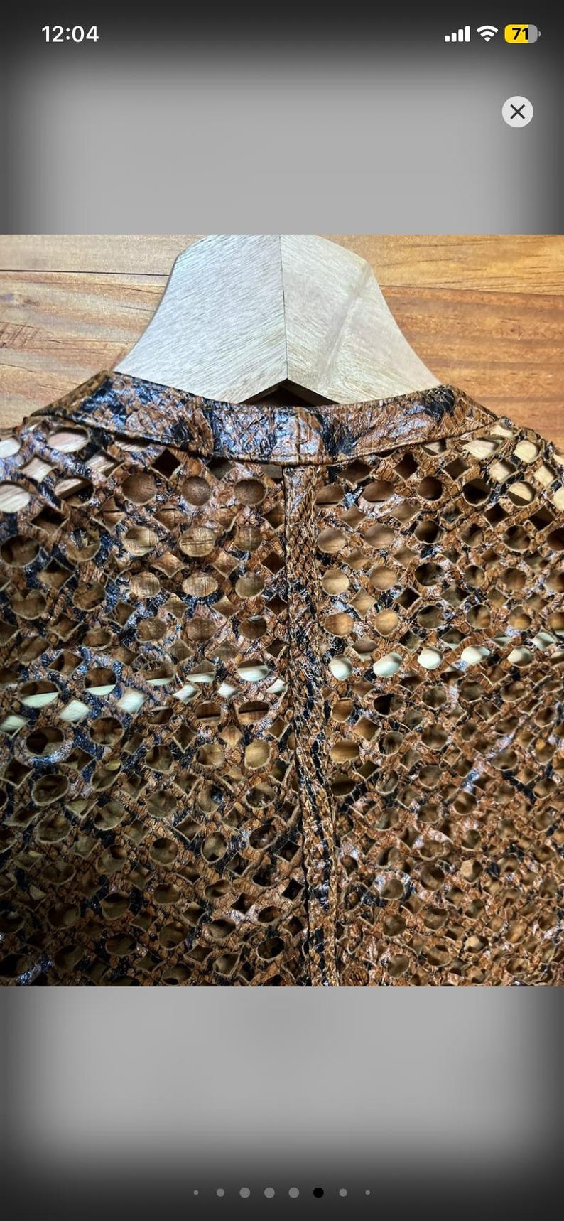 May include: Close-up of a brown and black open-weave jacket. The jacket has a unique pattern of cut-out shapes, creating an intricate design. The garment is displayed on a wooden hanger against a wooden background.