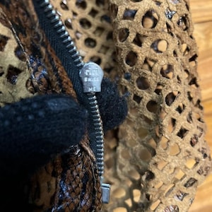 May include: Close-up of a brown and tan leather garment with a zipper. The zipper pull is silver and has the text "riri SWISS ME". The garment has a cut-out design and a snakeskin pattern.
