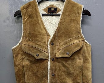 Op de afbeelding: Een bruin suède vest met een witte imitatiebontvoering. Het vest heeft een drukknoopsluiting en twee grote zakken aan de voorkant. Het vest is gelabeld met "Maverick" en "40".