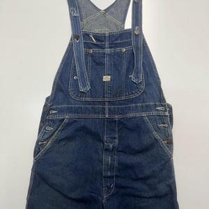 May include: Blue denim overalls with adjustable straps, a bib pocket, and side button closures. The overalls have multiple pockets and a classic workwear design. The denim is a medium wash, and the overall style is vintage.