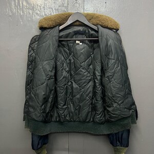 Puede incluir: Chaqueta bomber acolchada verde oscuro con cuello de piel marr&oacute;n, colgada de una percha de madera. La chaqueta tiene dobladillo y pu&ntilde;os de canal&eacute; y cierre de cremallera. El forro interior tambi&eacute;n es verde oscuro.