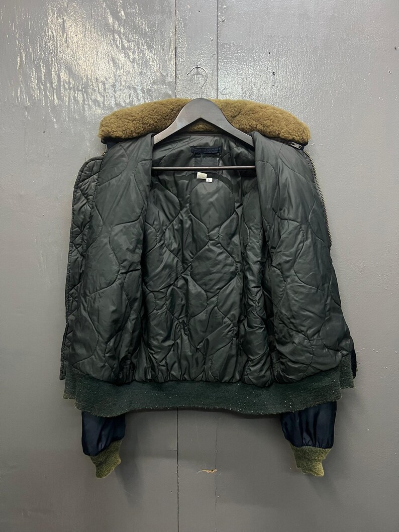 Puede incluir: Chaqueta bomber acolchada verde oscuro con cuello de piel marr&oacute;n, colgada de una percha de madera. La chaqueta tiene dobladillo y pu&ntilde;os de canal&eacute; y cierre de cremallera. El forro interior tambi&eacute;n es verde oscuro.