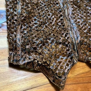 May include: Close-up of a brown, open-weave garment with a textured surface. The garment has a zipper and is made of a material that resembles snakeskin. The item is likely a vest or jacket.