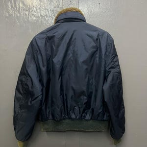 Puede incluir: Chaqueta bomber azul marino con cuello de piel marr&oacute;n y pu&ntilde;os y dobladillo de canal&eacute; verde. La chaqueta est&aacute; hecha de un material brillante. La parte trasera de la chaqueta es visible.