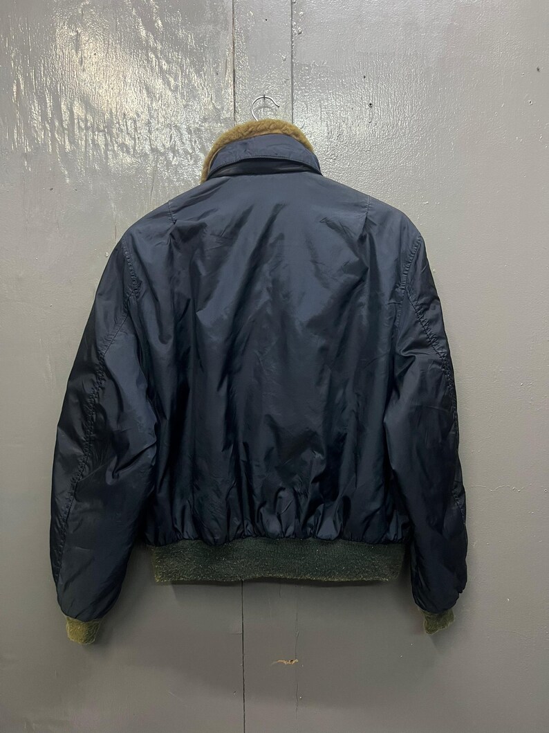 Puede incluir: Chaqueta bomber azul marino con cuello de piel marr&oacute;n y pu&ntilde;os y dobladillo de canal&eacute; verde. La chaqueta est&aacute; hecha de un material brillante. La parte trasera de la chaqueta es visible.