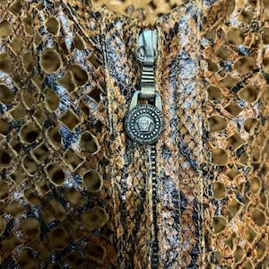 May include: Close-up of a brown and black patterned jacket with a zipper. The jacket has a unique design with cut-out details. The zipper pull features a decorative emblem.