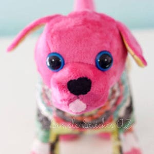 May include: A pink plush dog toy with blue eyes and a black nose. The dog is wearing a colourful patchwork fabric outfit.