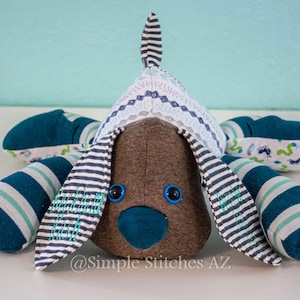 May include: A plush toy dog with blue and white striped ears and legs. The dog has a brown head with blue eyes and a blue nose. The dog is wearing a white and blue patterned hat. The dog is sitting on a white surface. @Simple Stitches AZ