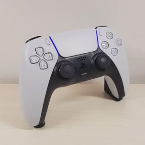 May include: A white and black PlayStation 5 DualSense wireless controller with a blue light bar. The controller has a touchpad, two analog sticks, and a D-pad.