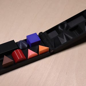 May include: A black, hinged, geometric shape sorter with various colorful shapes. The shapes include a blue cube, red cylinder, orange triangle, and a black rectangle. The sorter is open, revealing compartments for each shape.