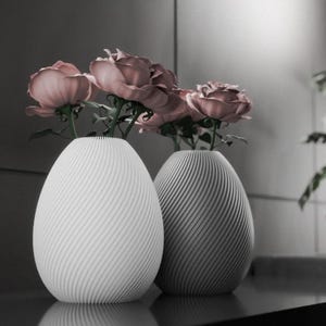 Pode incluir: Dois vasos de cerâmica branca com um padrão em espiral, um com um acabamento cinza claro e o outro com um acabamento branco, contendo rosas cor-de-rosa.