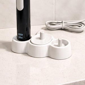 Könnte beinhalten: Eine schwarze elektrische Zahnbürste mit gelben Borsten und eine weiße Ladestation. Ein weißes Nachtlicht ist in eine schwarze Steckdose gesteckt. Die Zahnbürste steht neben einer kleinen Topfpflanze.