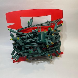 May include: A red plastic Christmas light holder with a bundle of green Christmas lights. The lights have small, colorful bulbs and are wrapped around the holder. The holder has a rectangular shape.