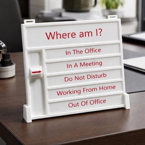 May include: A white office status sign with red text. The sign reads "Where am I?" with options including "In The Office," "In A Meeting," and "Do Not Disturb." A red arrow indicates the current status. The sign is on a brown desk.