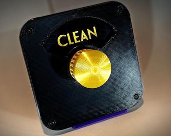 Könnte beinhalten: Ein schwarzer quadratischer Knopf mit einem goldenen Knopf und einem weißen Etikett mit der Aufschrift "CLEAN".
