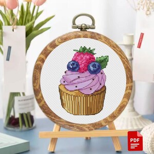 Peut inclure: Un motif de point de croix représentant un cupcake avec un glaçage violet et une framboise et des myrtilles sur le dessus. Le motif est encadré dans un cadre rond en bois et exposé sur un petit chevalet en bois.