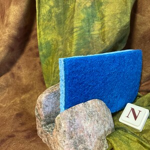 May include: A blue sponge sits in a stone holder with a letter N tile in front of it. The holder is made of rough, gray stone with brown and white streaks. The background is a green and brown fabric.