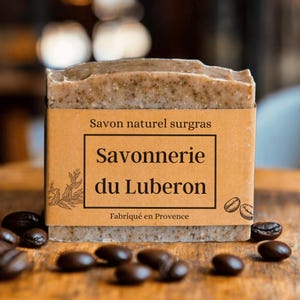 Könnte beinhalten: Ein rechteckiger Block Naturseife mit einem braunen Papieretikett. Auf dem Etikett steht "Savonnerie du Luberon" und "Fabriqué en Provence". Kaffeebohnen sind um die Seife auf einer Holzoberfläche verstreut.