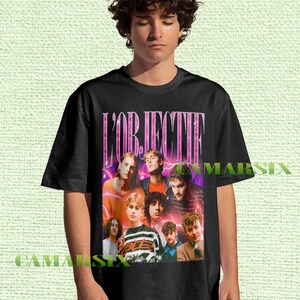 Może przedstawiać: Czarny t-shirt z grupowym zdjęciem i różowym napisem "L'OBJECTIF". Projekt ma estetykę retro, z fioletowymi i pomarańczowymi odcieniami. Koszulka jest noszona przez osobę.