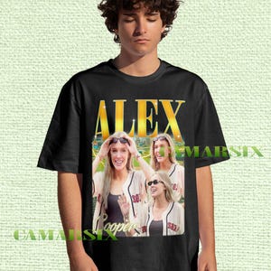 Puede incluir: Camiseta negra con un gráfico de tres mujeres con gafas de sol y camisetas de béisbol. La palabra "ALEX" está impresa en oro encima de la imagen, con "Cooper" debajo. La camiseta la lleva una persona.
