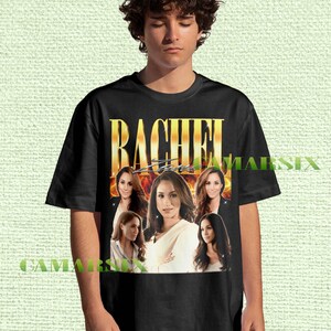 May include: Black t-shirt featuring the name "RACHEL" in gold lettering above a collage of images of a woman. The shirt has a vintage, retro design.