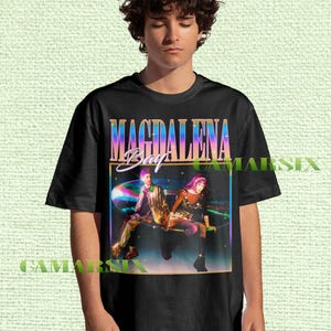 May include: Black t-shirt featuring the band name "MAGDALENA Bay" in a colorful, retro font. The graphic includes a vibrant image of two people in a futuristic setting. The shirt is a classic crew neck style.