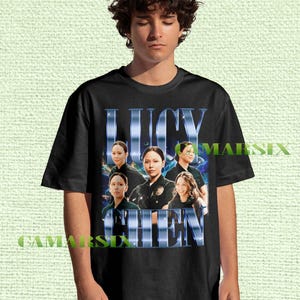May include: Black t-shirt featuring the name "LUCY CHEN" in a blue, stylized font. The shirt displays a collage of images of a person. The shirt is worn by a person.