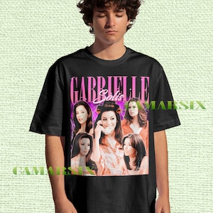 Puede incluir: Camiseta negra con un diseño gráfico que presenta al personaje de Gabrielle Solis de la serie Desperate Housewives. El diseño incluye el nombre "Gabrielle Solis" en rosa y varias imágenes del personaje.
