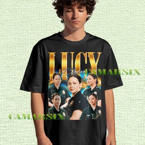 May include: Black t-shirt featuring a gold-colored "LUCY" text design with a portrait of a woman in a police uniform. The shirt also includes the text "Chen" and multiple images of the same woman.