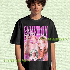 May include: Black t-shirt featuring a retro design with the name "CAMERON Diaz" in a pink, vertical font. The shirt includes multiple images of the actress. The shirt is worn by a person.
