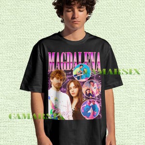 May include: Black t-shirt featuring the name "MAGDALENA" in a pink and purple retro font. The shirt includes images of a musical group and the word "Bay". The shirt is worn by a person.