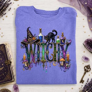 Può includere: Una t-shirt girocollo viola con una stampa grafica della parola WITCHY in grandi lettere ornate e multicolori. Il design include un gatto nero, un cappello da strega, boccette di pozione in vetro, funghi e candele in stile decorativo.