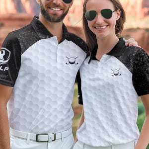 May include: Two people wearing white golf shirts with a golf ball pattern and black sleeves. The shirts have a small golf club and ball logo. They are also wearing white pants and sunglasses. The background features a scenic view of red rock formations and green grass.