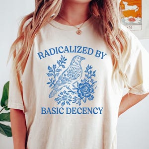 Puede incluir: Camiseta de color marfil con el texto azul "RADICALIZED BY BASIC DECENCY". Un diseño de pájaro y flores azules está centrado en la camiseta. La camiseta tiene un corte relajado.